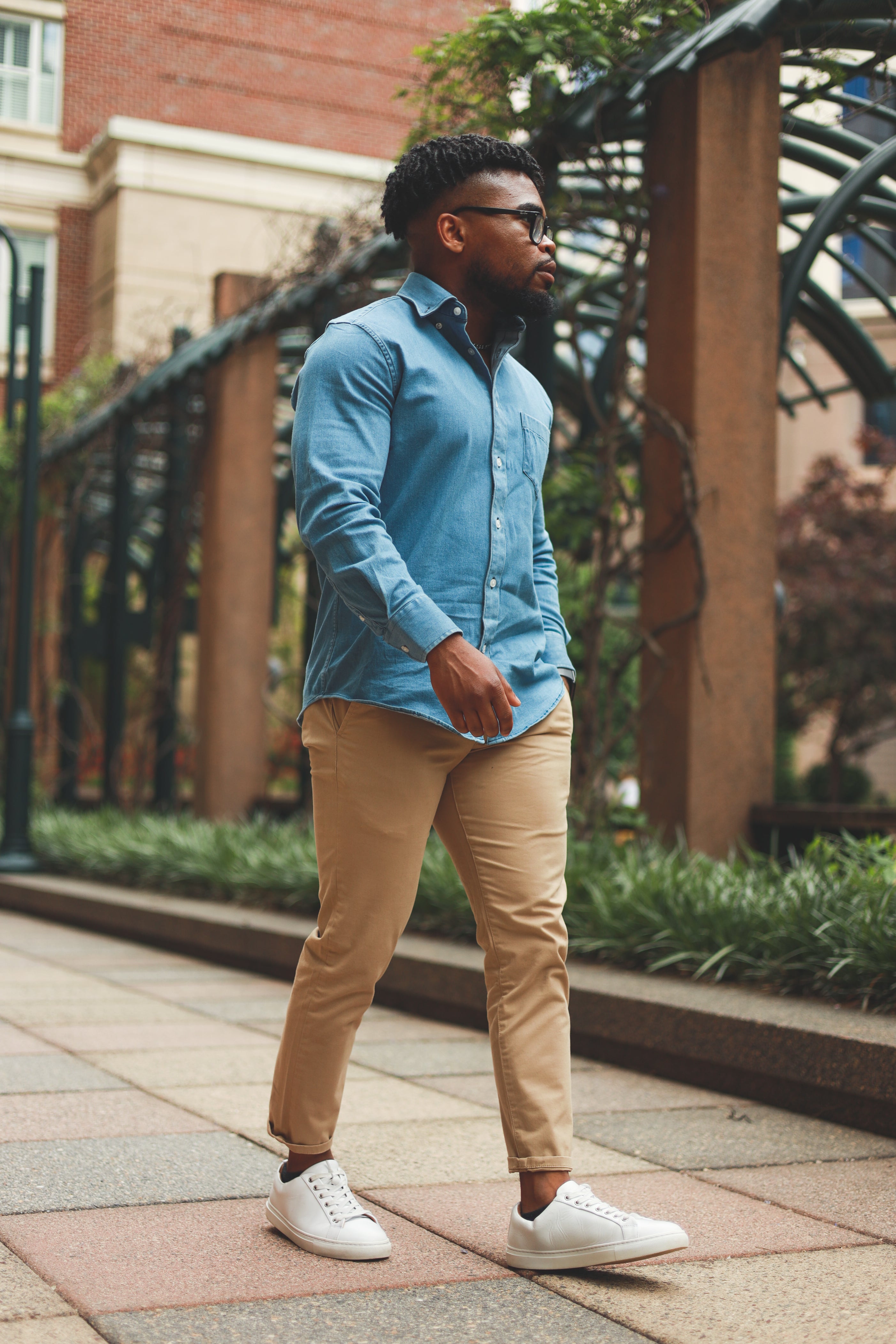 This image depicts a stylish man walking outdoors. He is wearing a light blue button-down shirt, beige chinos, and white sneakers. The setting appears to be an urban area with greenery and a brick building in the background. The outfit is casual yet polished, suitable for various occasions.