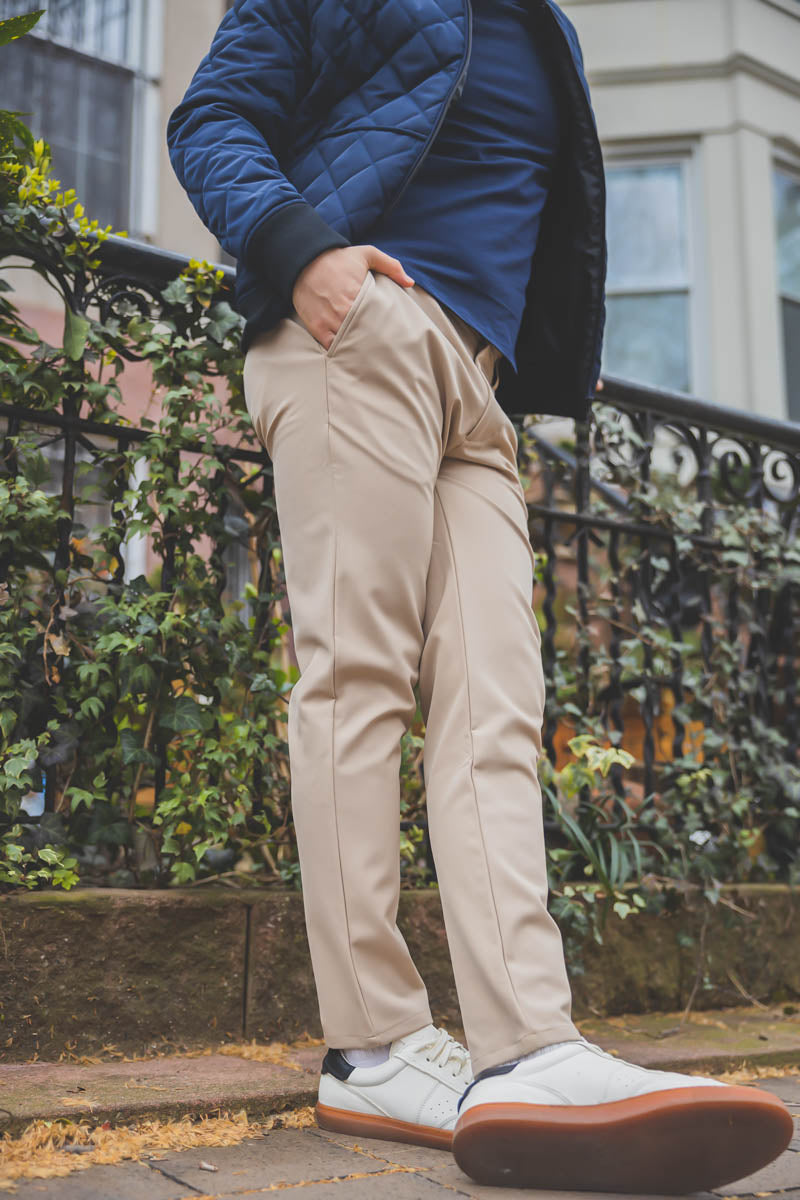 The image shows a person standing outdoors, wearing beige pants and white sneakers with brown soles. The person is also wearing a dark blue quilted jacket and a navy blue shirt. They have their hands in their pockets, and the background includes a wrought iron fence with greenery climbing on it, along with a building partially visible in the distance. The setting appears to be a residential or urban area.